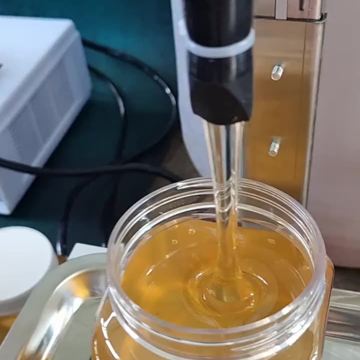 Close-up of anti-drip honey filling nozzle dispensing a smooth stream into a wide-mouth jar on the weighing tray